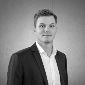 Professionelles Porträt eines jungen Mannes in einem Anzug mit Hemd, vor einem neutralen, hellen Hintergrund. Ideal für geschäftliche Profile oder Unternehmenswebsites.